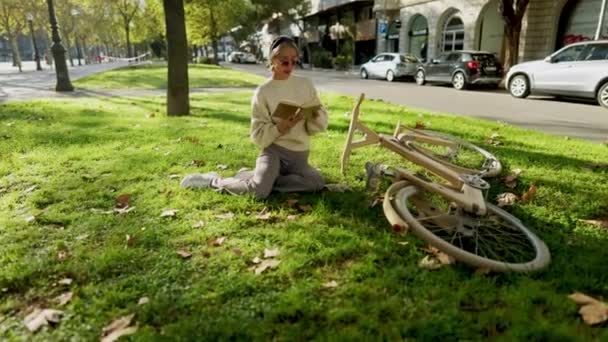 Mujer leyendo un libro en el parque en el césped. tiene bicicleta ecológica de madera — Vídeos de Stock