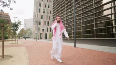 Açık havada akıllı telefonları olan Suudi bir adam, yolda yürürken telefonla konuşan Arap bir işadamı.