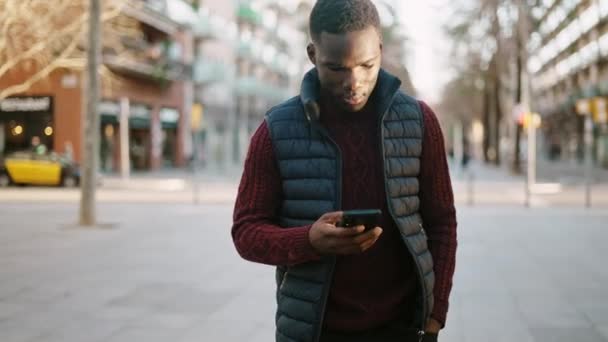 Hombre negro navegando teléfono inteligente y caminando en la ciudad — Vídeo de stock