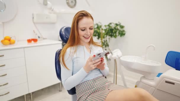 Jovem sorrindo mulher em cadeira dental usando telefone celular enquanto espera por ortodontista na clínica — Vídeo de Stock