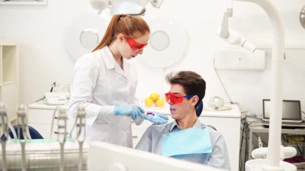 Молодая женщина-стоматолог в ультрафиолетовых защитных очках с помощью света для стоматологического лечения во время композитного заполнения — стоковое видео
