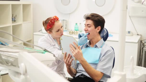 Профессиональная женщина-стоматолог держит зеркало и показывает результат лечения зубов мужчине — стоковое видео