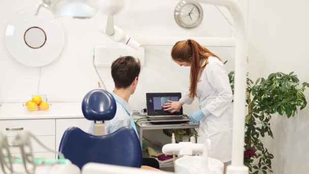 Женщина-стоматолог и мужчина смотрят на ноутбук с помощью рентгена и обсуждают план лечения зубов — стоковое видео