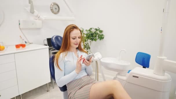 Joven paciente femenina positiva sentada en silla dental con teléfono inteligente en la oficina del dentista moderno — Vídeo de stock
