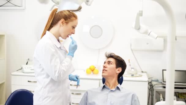 Молода дружня жінка-стоматолог розмовляє з пацієнтом чоловічої статі, сидячи в кріслі про гігієну здоров'я порожнини рота — стокове відео