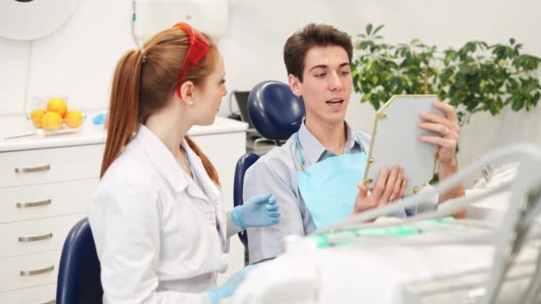Dentista femenina sosteniendo espejo y mostrando problemas dentales a paciente masculino en consultorio dental — Vídeos de Stock