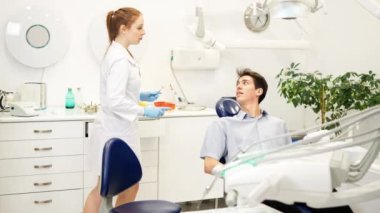 Kadın dişçi, klinikteki diş kontrolü sırasında aynalı genç adamın dişlerini inceliyor.