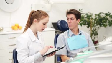 Genç dişçi dijital tablete bakıyor ve dişçi kliniğindeki erkek hastayı dinliyor.