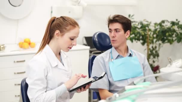 Jeune dentiste féminine regardant tablette numérique et écoute patient masculin en clinique dentaire — Video