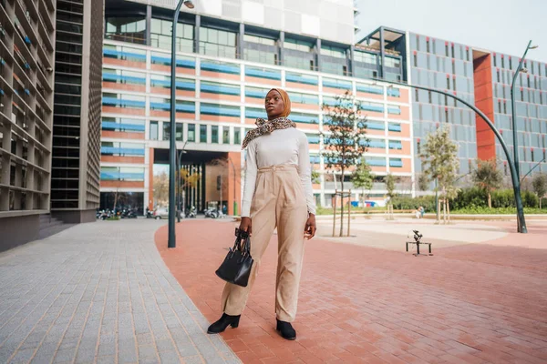 Mujer negra en ropa casual inteligente y hijab de pie cerca de rascacielos — Foto de Stock