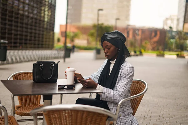 Mujer negra seria usando teléfono inteligente en la mesa en la cafetería al aire libre —  Fotos de Stock