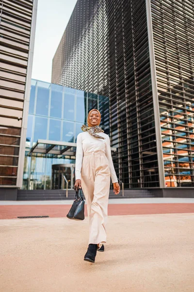 Mujer negra en ropa casual inteligente y hijab de pie cerca de rascacielos —  Fotos de Stock