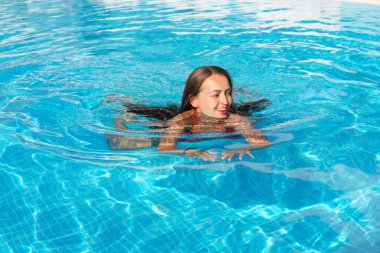 Yüksek açılı, uzun siyah saçlı, neşeli genç bir kadın. Güneşli bir hafta sonu, temiz havuzda yüzerken gülümseyip başka tarafa bakıyor.