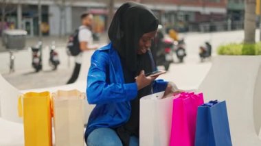 Moda elbiseli ve tesettürlü Müslüman bir kadın alışveriş sonrası kenarda otururken renkli kağıt torbalarla malların fotoğraflarını çekiyor.