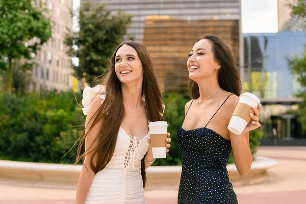 Delighted young female friends in dresses with paper cups of takeaway coffee standing in street and enjoying weekend in summer