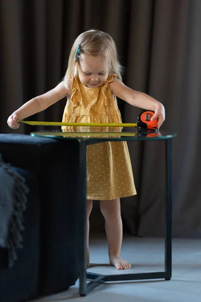 A little two-year-old girl measures the length of the table with a tape measure —  Fotos de Stock