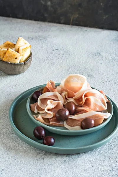 Slices of thinly sliced ham (prosciutto, Parma ham) with a bowl of olives on a light gray background. An appetizing Mediterranean snack, antipasti.