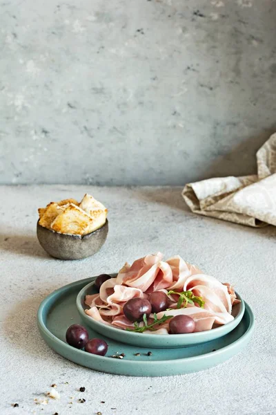 Slices of thinly sliced ham (prosciutto, Parma ham) with a bowl of olives on a light gray background. An appetizing Mediterranean snack, antipasti.