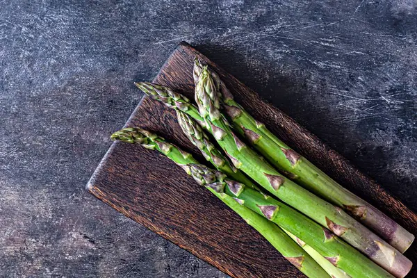 Koyu arkaplandaki tahta kesme tahtasındaki yeşil kuşkonmaz. Çiğ gıda konsepti, kopyalama alanı.