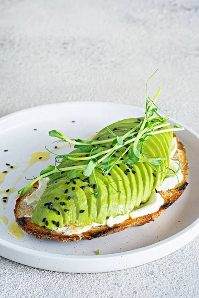 Whole grain avocado toast with cream cheese, black sesame seeds, olive oil and microgreens (sprouted pea seeds) on a light background. Healthy food concept.
