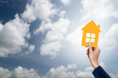 Mavi güneşli gökyüzü arka planında sarı kağıt ev tutuyordu. Bir ev satın alma görüntüsü.