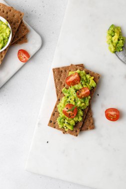 Ekmeğe avokado ezmeli, domatesli, kirazlı ve roka beyaz masada. Kapat.