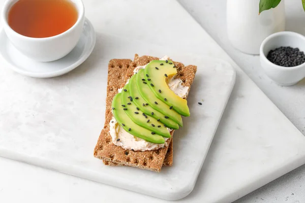 Avokado tostlu kahvaltı, mermer tahtada siyah susam tohumlu tam ekmekli. Bir fincan çay..