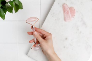 Kadın elinde pembe tekerlekli masaj aleti ve fayans arka planında da guaj kazıyıcısı. Yaş karşıtı, ürün kaldırma.