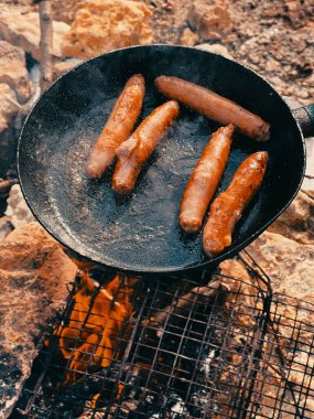 Sosisler kızartma tavasında kızartılır. Piknik. Turist yemeği. Piknik. Fastfood. Izgarada ızgara sosis, duman ve alevle.