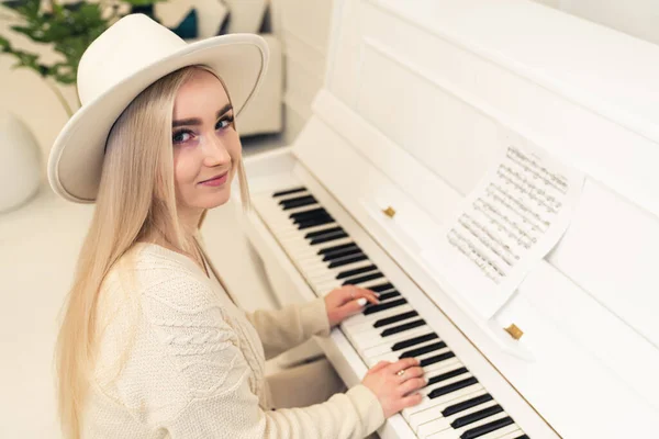 Femme européenne blonde compositeur aux cheveux longs dans un chapeau assis, jouant du piano, et regardant la caméra — Photo