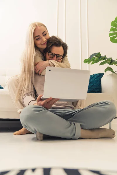 Вертикальный снимок милой кавказской пары, сидящей на полу и счастливо смотрящей на экран ноутбука и просматривающей интернет — стоковое фото