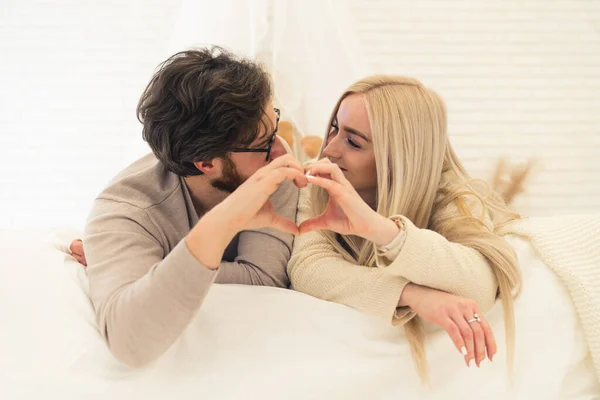 Bonito casal caucasiano olhando um para o outro formando forma de coração com as palmas das mãos enquanto deitado em sua cama — Fotografia de Stock