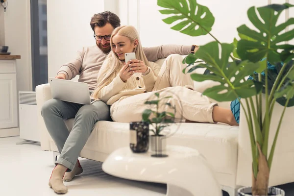 Schattig jong volwassenen paar met laptop en smartphone zitten op witte bank en genieten van netwerken en sociale media — Stockfoto