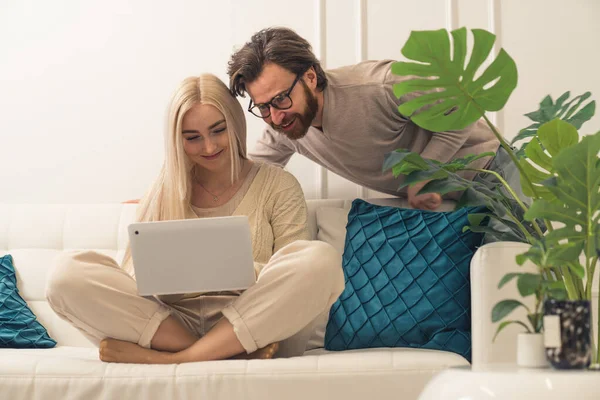 Aantrekkelijke vrolijke getrouwde echtgenoten die op een bank zitten, cadeautjes kopen en online winkelen dankzij hun gloednieuwe laptop — Stockfoto