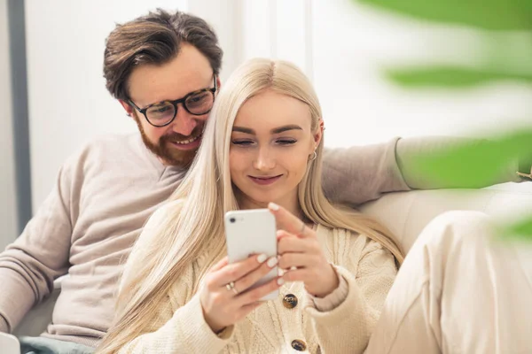 Europees duizendjarig echtpaar kijkt vrolijk naar een smartphone terwijl ze samen op een bank zitten — Stockfoto