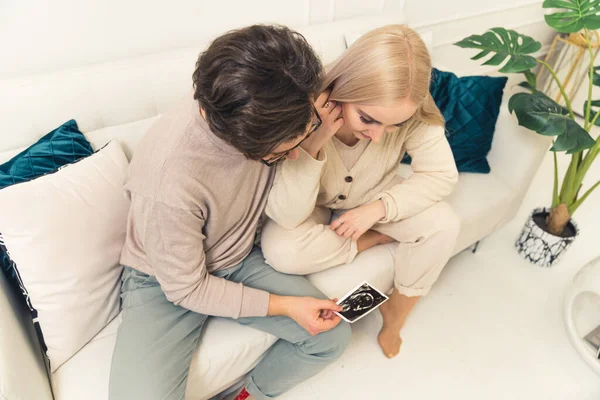 Genç beyaz bir çift, modern dairelerinde beyaz bir kanepede otururken ultrason fotoğraflarına bakıyor. — Stok fotoğraf