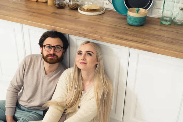 Прекрасна біла пара сидить на підлозі на сучасній кухні і дивиться вгору, думаючи про майбутнє — стокове фото