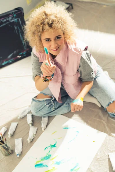 Joven artista femenina pintando la obra con pintura acrílica - señalando con el dedo a la cámara —  Fotos de Stock