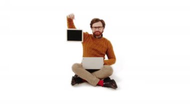 Kafkasyalı orta yaşlı girişimci dizlerinin üzerinde dizüstü bilgisayarla yan yana oturuyor ve siyah ekranlı bir tablet beyaz arka plan çekimleri yapıyor.