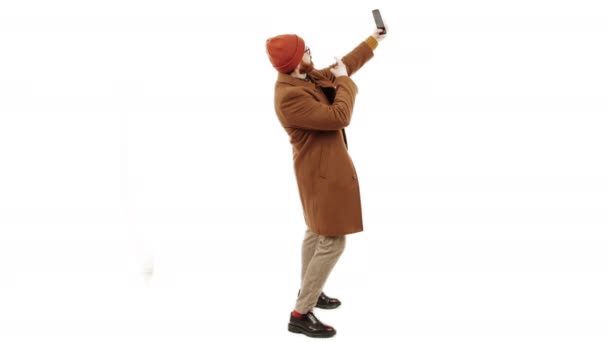 Uomo europeo in un cappotto e un berretto scattare un selfie o in streaming tramite smartphone isolato studio girato sfondo bianco orizzontale — Video Stock