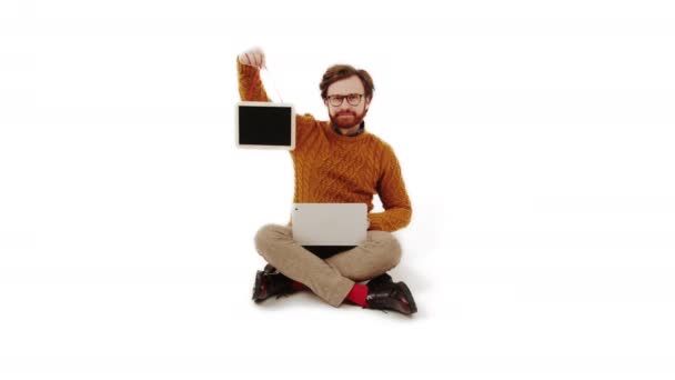 Кавказский предприниматель средних лет, сидящий со скрещенными ногами с ноутбуком на коленях и планшетом с черным экраном на белом фоне, снятый студией — стоковое видео