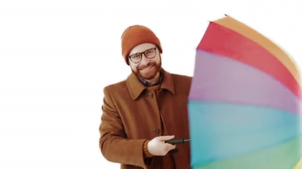 Jeune homme beau avec barbe vêtu d'imperméable pour la journée de pluie tenant parapluie arc-en-ciel coloré, le tournant autour, et souriant en regardant la caméra, studio isolé tourné fond blanc — Video
