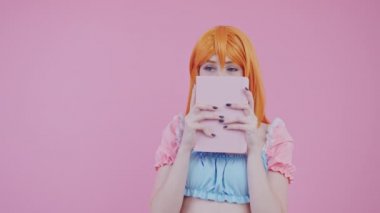 Utangaç, kızıl saçlı, yüzünü örten ve arka plandaki pembe stüdyo fotoğrafının arkasına bakan bir kadın yazar.