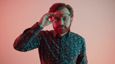 Yakışıklı beyaz sakallı adam somon pembe arka plan çekiminin başında dikilirken tek eliyle modern gözlüklerine dokunuyor.