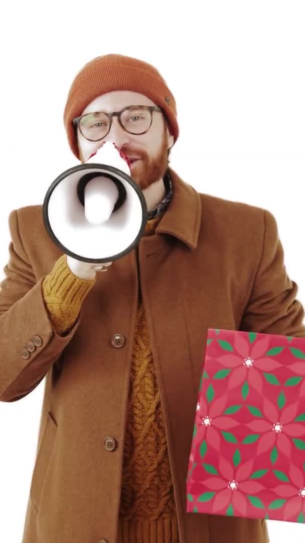 Hombre caucásico inteligente en un abrigo y gorro mirando a la cámara anunciando grandes ventas a través de altavoz y sosteniendo bolsa de papel estudio vertical plano fondo blanco — Vídeo de stock