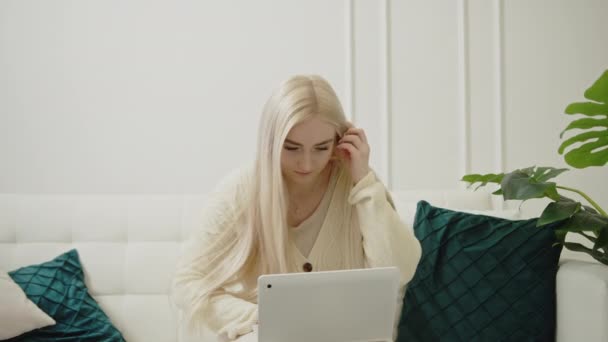 Європейська молода блондинка читає статтю онлайн через свій ноутбук сидячи на дивані в своїй сучасній квартирі. — стокове відео