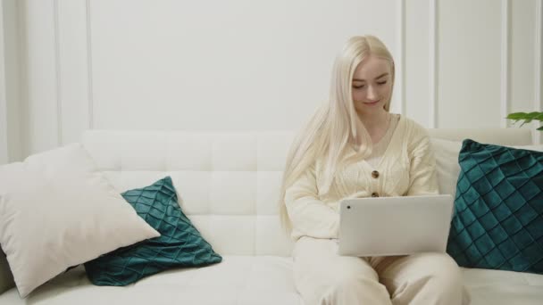 Αρκετά καυκάσια νεαρή γυναίκα που εργάζεται με φορητό υπολογιστή κάθεται σε λευκό καναπέ στο σύγχρονο σαλόνι της — Αρχείο Βίντεο