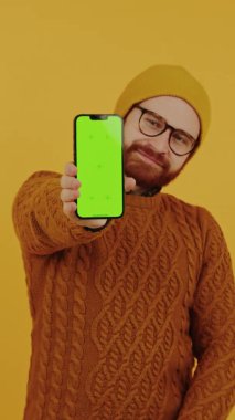 Beyaz adam yeşil ekran cep telefonu tutuyor ve sarı arkaplan üzerinde dikey çekimler yapıyor.