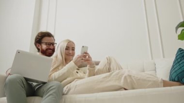 Modern Kafkasyalı genç yetişkinler bir kanepede dizüstü bilgisayar ve telefonla birlikte oturup internette geziniyorlar.