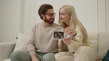 Neşeli bin yıllık Kafkasyalı kadın, kocasını kucaklıyor ve oturma odasındaki bir kanepede otururken gelecekteki bebeklerinin ultrason fotoğrafını tutuyor.